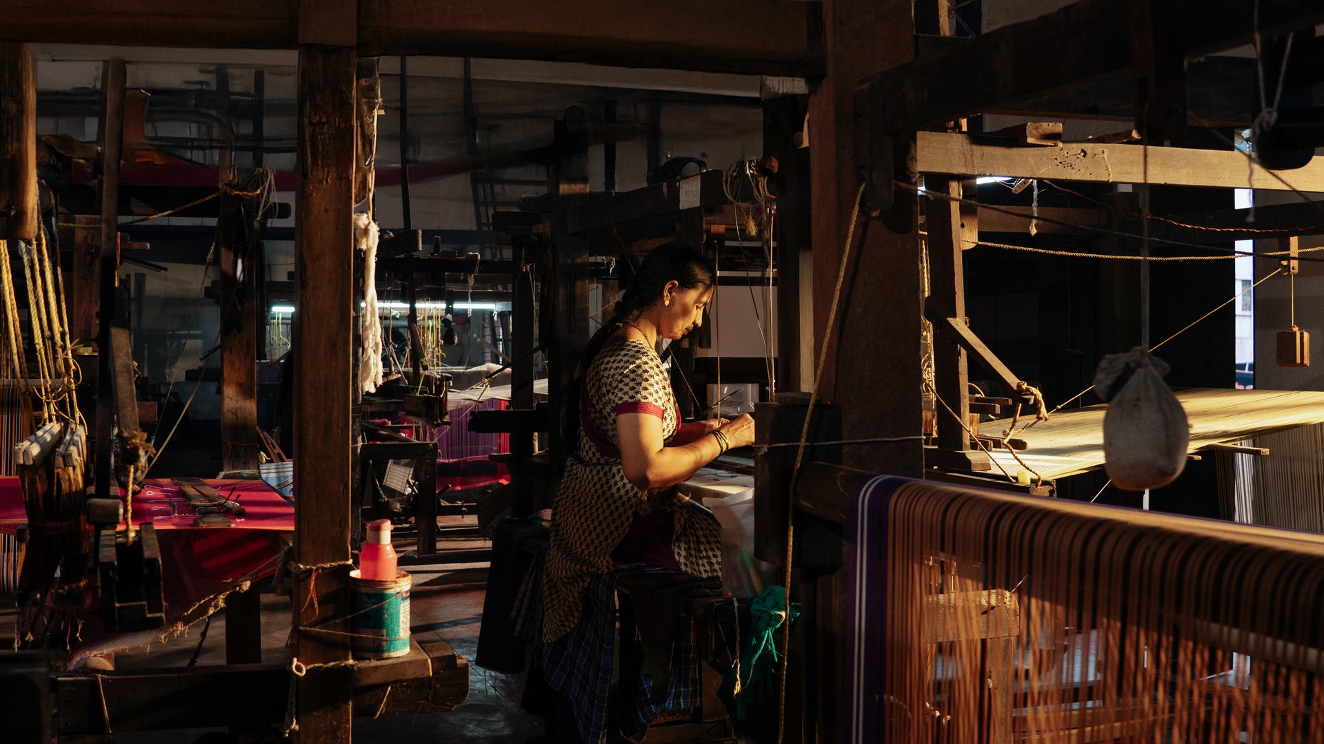 Person working on a loom in a dimly lit room with machinery and equipment.